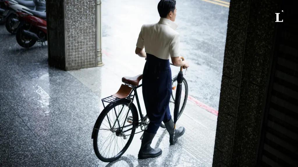 一名男性背對鏡頭，推著黑色腳踏車在濕滑的騎樓邊緣行走。他身穿米白色立領短袖外套，內搭深藍色長褲，搭配黑色高筒雨鞋。左手扶著腳踏車把手，右手握著一把黑色的摺疊傘，傘尖朝下滴水。地面因雨水而反光，可見騎樓柱子旁的濕滑地磚和背景處的雨景與停放的機車。